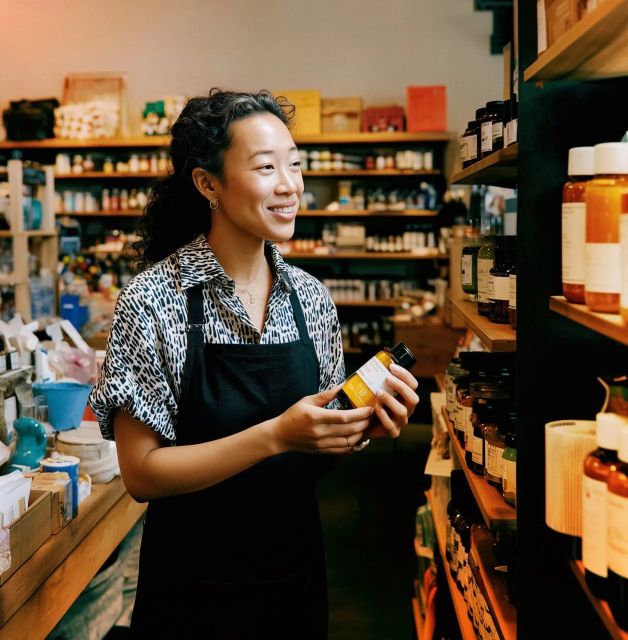 A smiling retail store associate in a black apron checking product inventory on wooden shelves in a boutique shop.