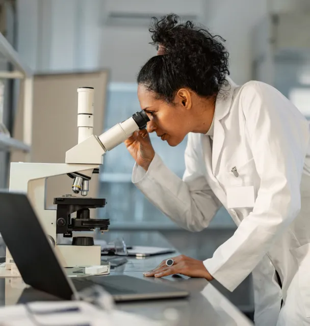 A female wearing a lab gown looks at a microscope beside a laptop