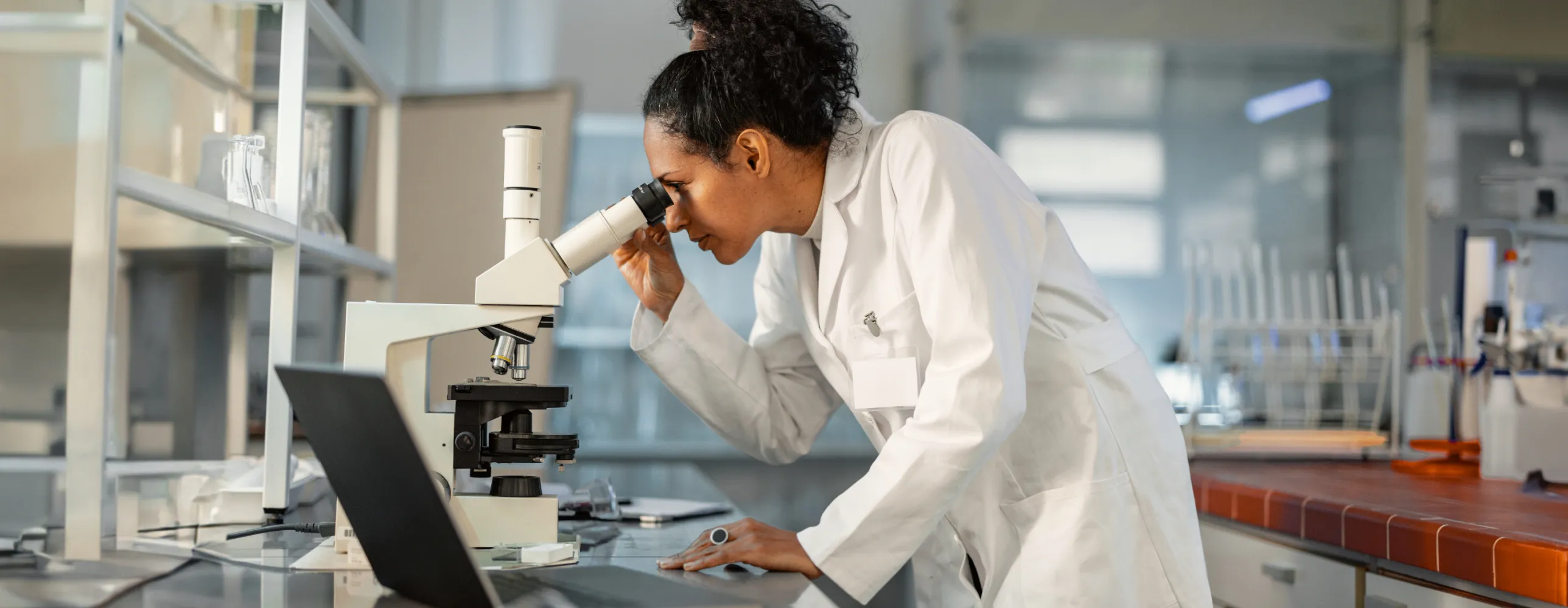 A female wearing a lab gown looks at a microscope beside a laptop